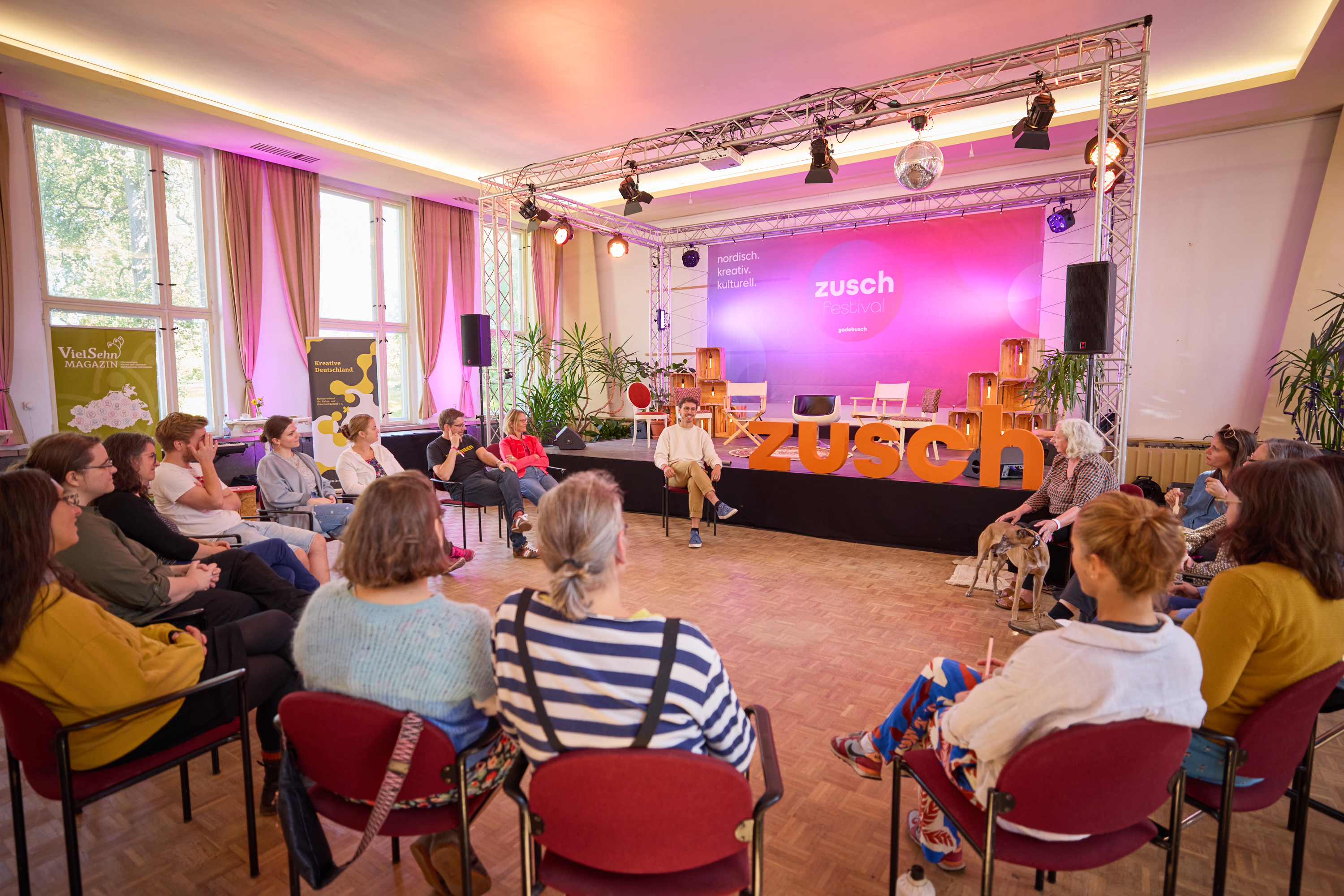 Menschen sitzen in einem Stuhlkreis in einem großen Saal vor der Bühne